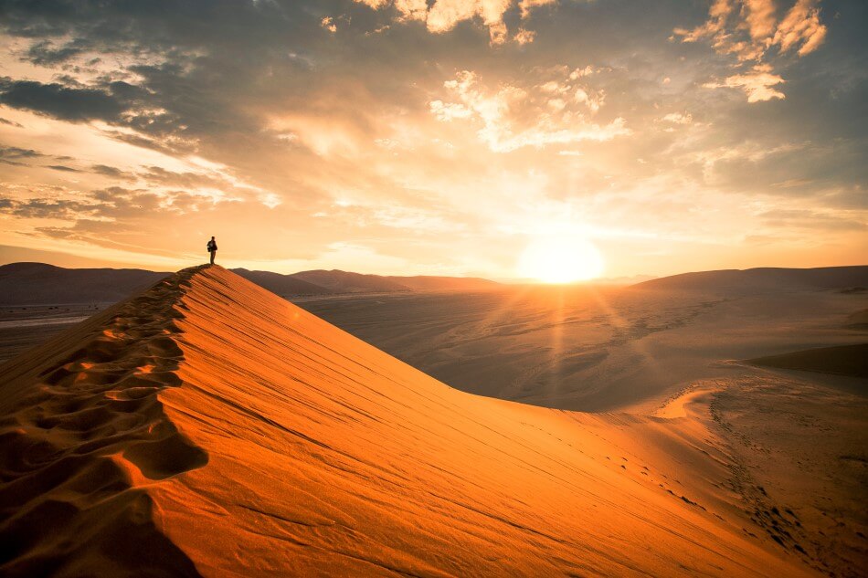 A person observes a sunrise while stood on top of a sand dune in the Namibian Desert.